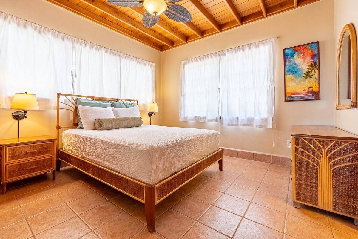 A bedroom with a wooden bed, two bedside tables, a ceiling fan, and a painting on the wall.