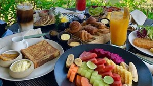 Une table de petit-déjeuner garnie de divers aliments et boissons, avec vue sur une plage bordée de palmiers et sur l’océan.