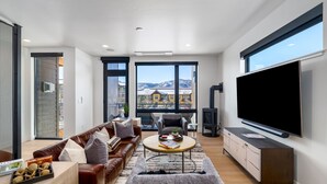 A modern living room with a large TV, leather sofas, a coffee table, and a view of mountains through large windows. - Urban Reserve, AC! Upscale, Private Rooftop Hot Tub & Deck Area with Views! (Steamboat Springs)