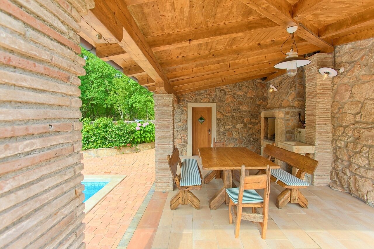 Eine überdachte Terrasse mit einem Holztisch und Bänken, Steinwänden und Blick ins Grüne.