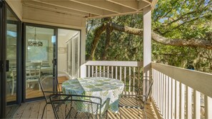 Un espace extérieur abrité, doté d’une table et de chaises, d’une porte vitrée donnant sur l’intérieur, et offrant une vue sur les arbres et le ciel.