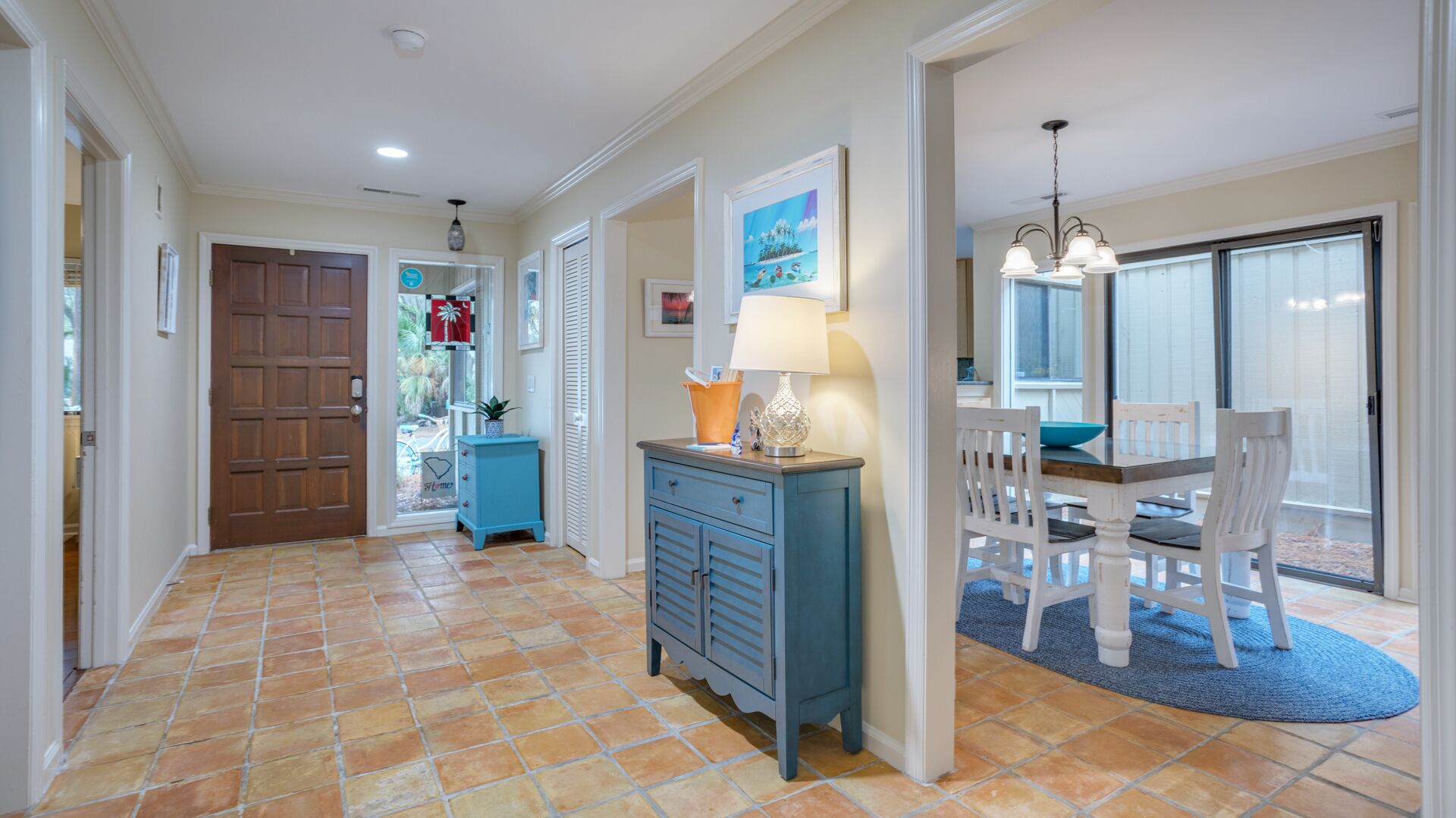 A hallway with a tiled floor, a wooden door, a blue cabinet, a table with chairs, and a window with blinds.
