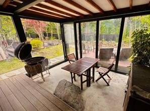 Eine überdachte Terrasse mit Holztisch und -stühlen, einem Grill und Blick auf einen Garten mit Bäumen und einem Zaun.