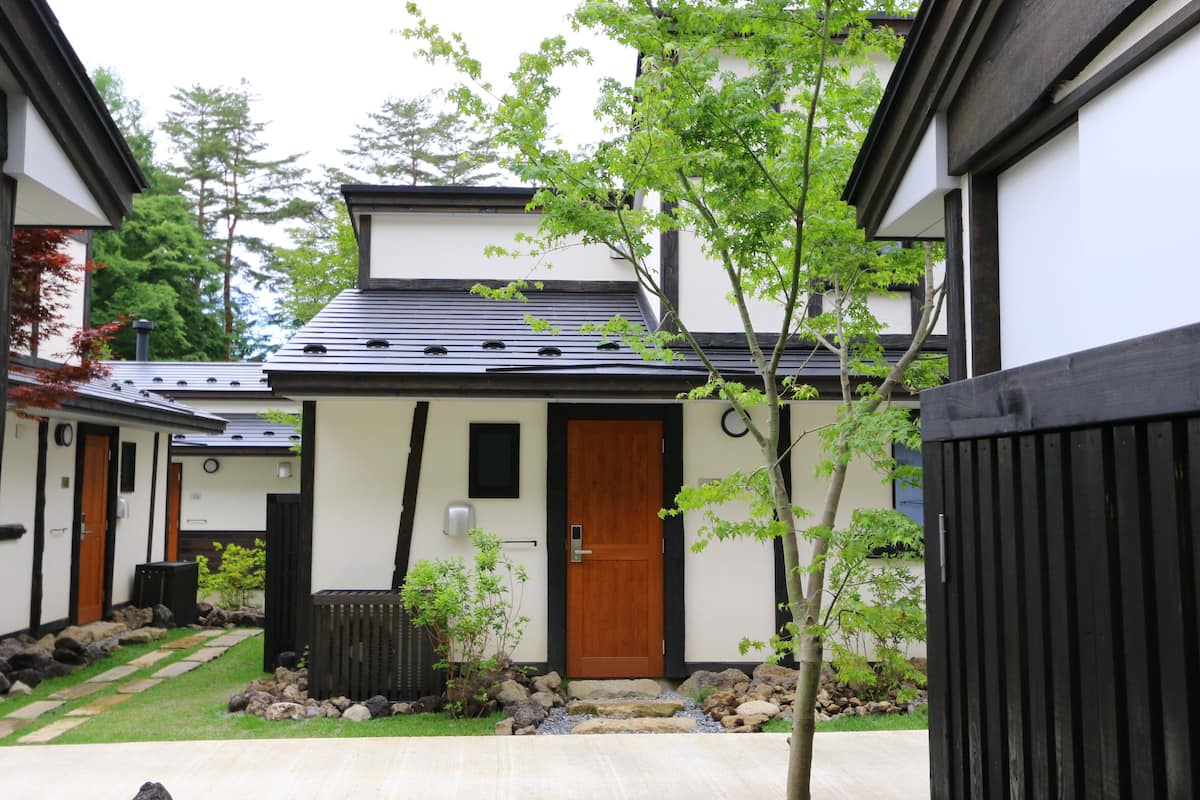 Un bâtiment de style japonais traditionnel, doté d’une porte en bois, d’un petit jardin agrémenté de pierres plates servant de pas et d’une clôture noire.