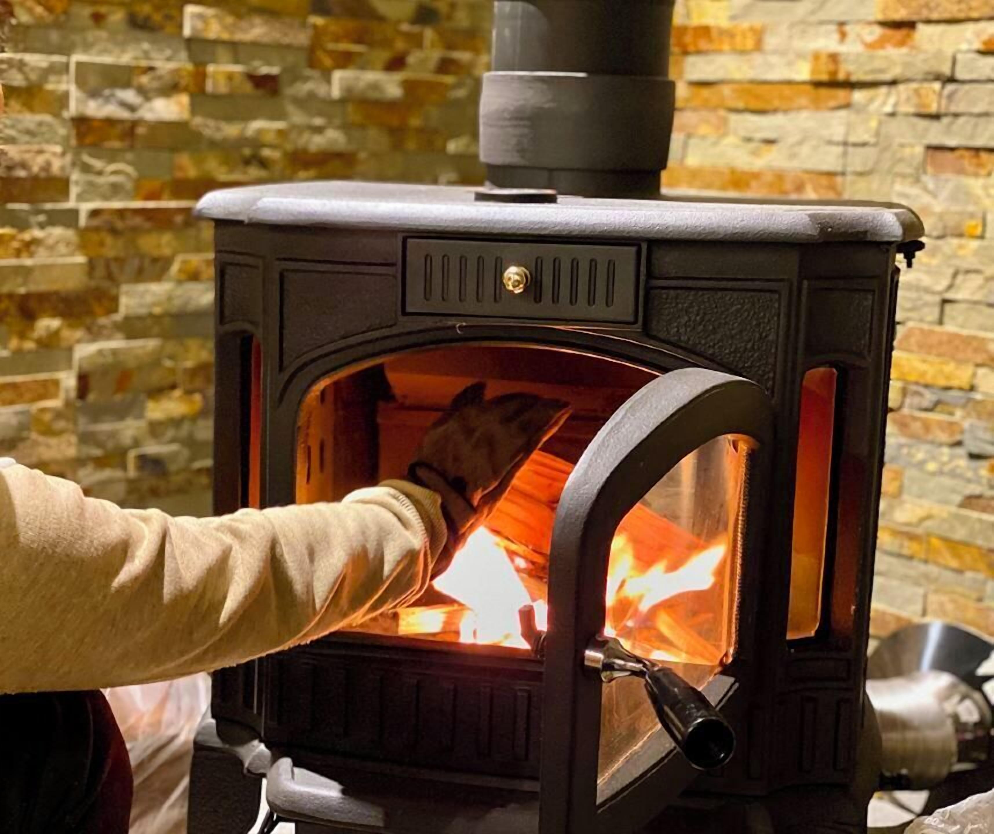 A wood-burning stove with a glass door, a person adjusting the fire, and a stone wall background.