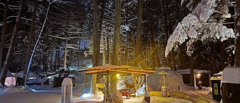 A snow-covered outdoor area with a wooden gazebo, benches, and a trash can.