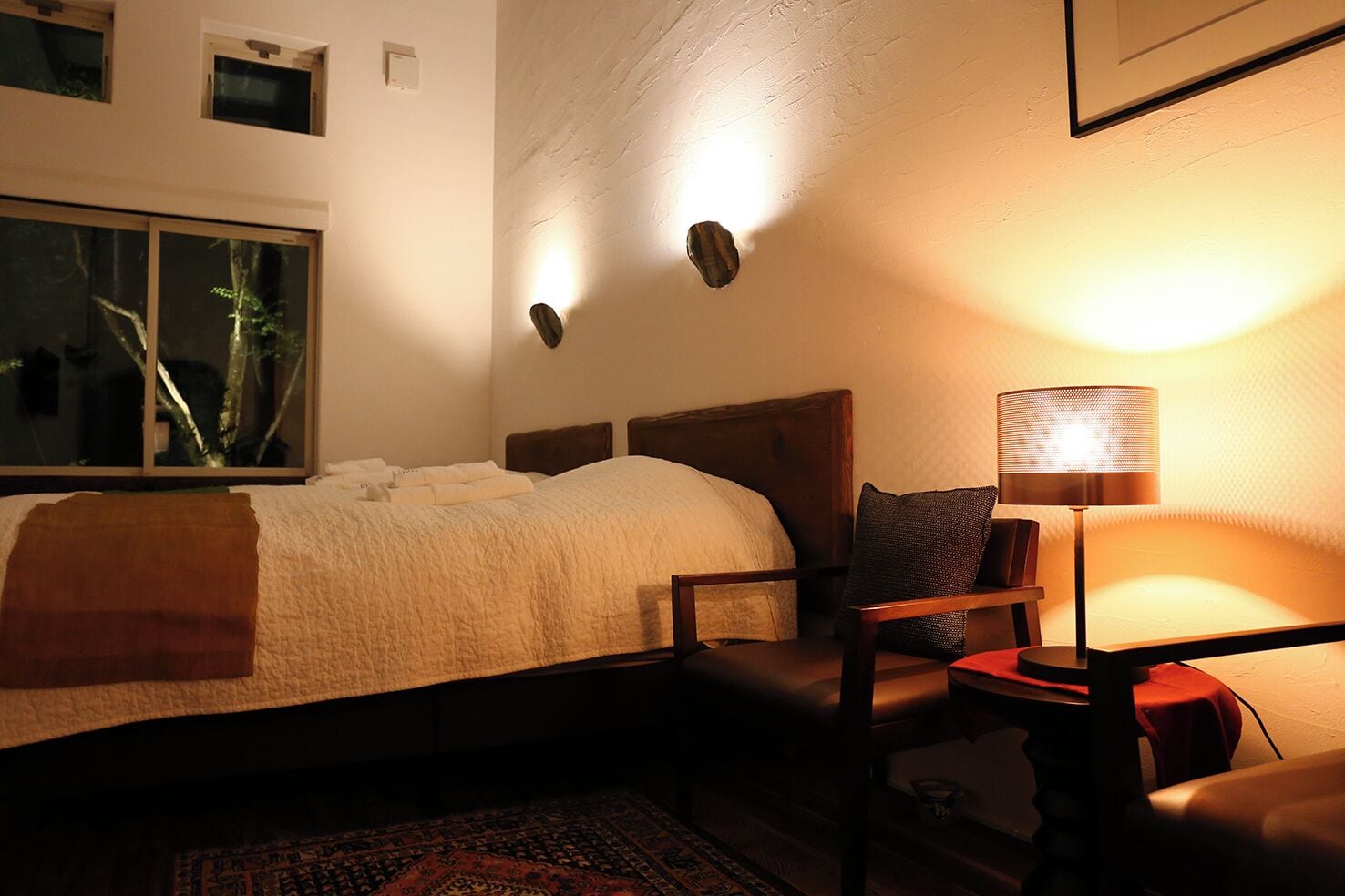 A hotel room with a bed, a chair, a table, a lamp, and a window with a view of trees.