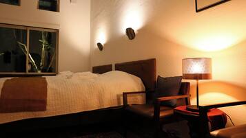 Ein Hotelzimmer mit Bett, Sessel, Tisch, Lampe und Fenster mit Blick auf Bäume.