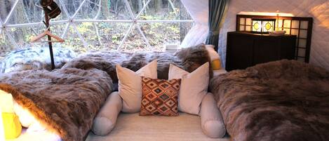 A cozy tented room with a large window, a bed with a furry blanket, and a bedside table with a lamp.