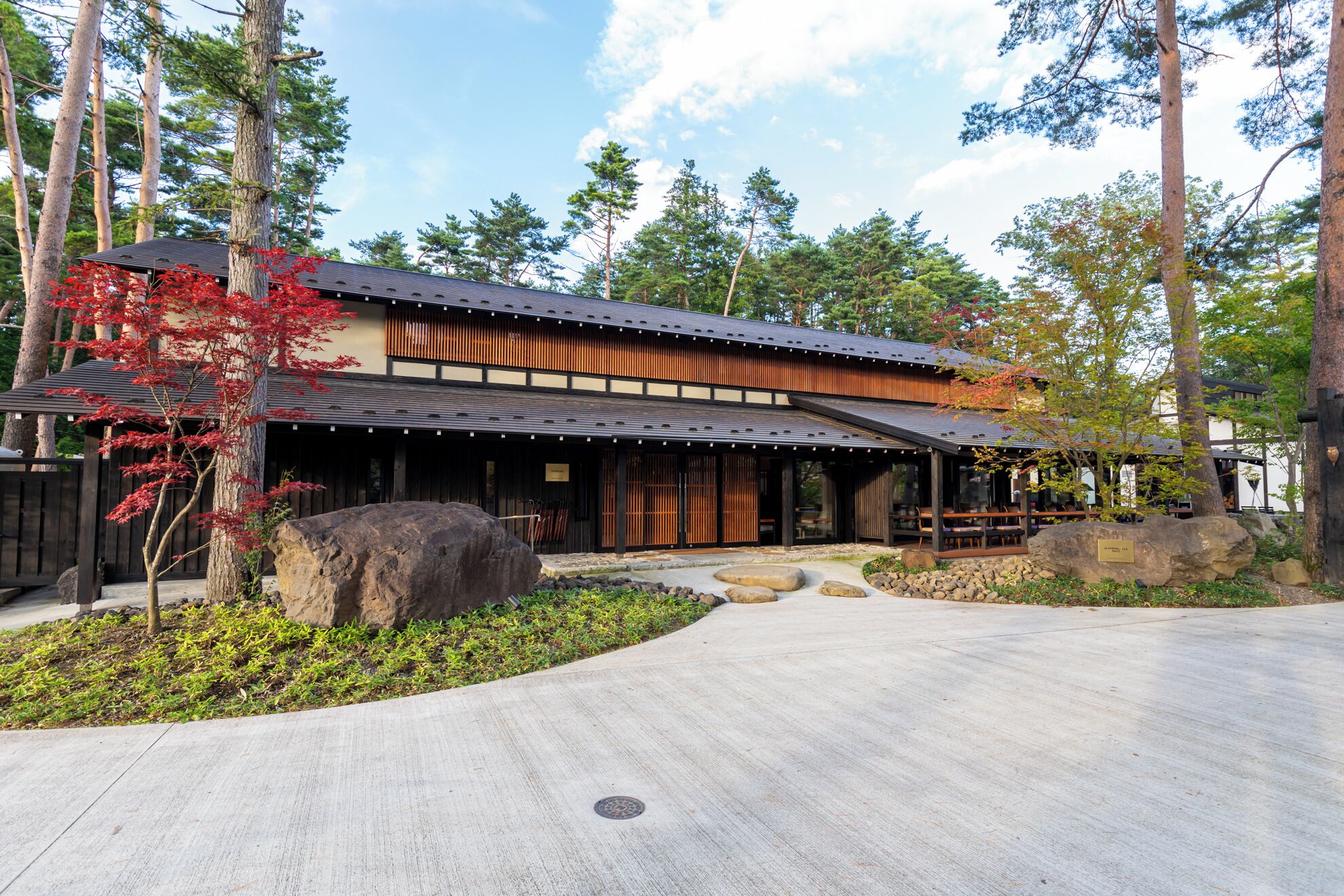 Un edificio tradicional japonés con fachada de madera y tejado de tejas, rodeado de un jardín ajardinado con rocas grandes y árboles.