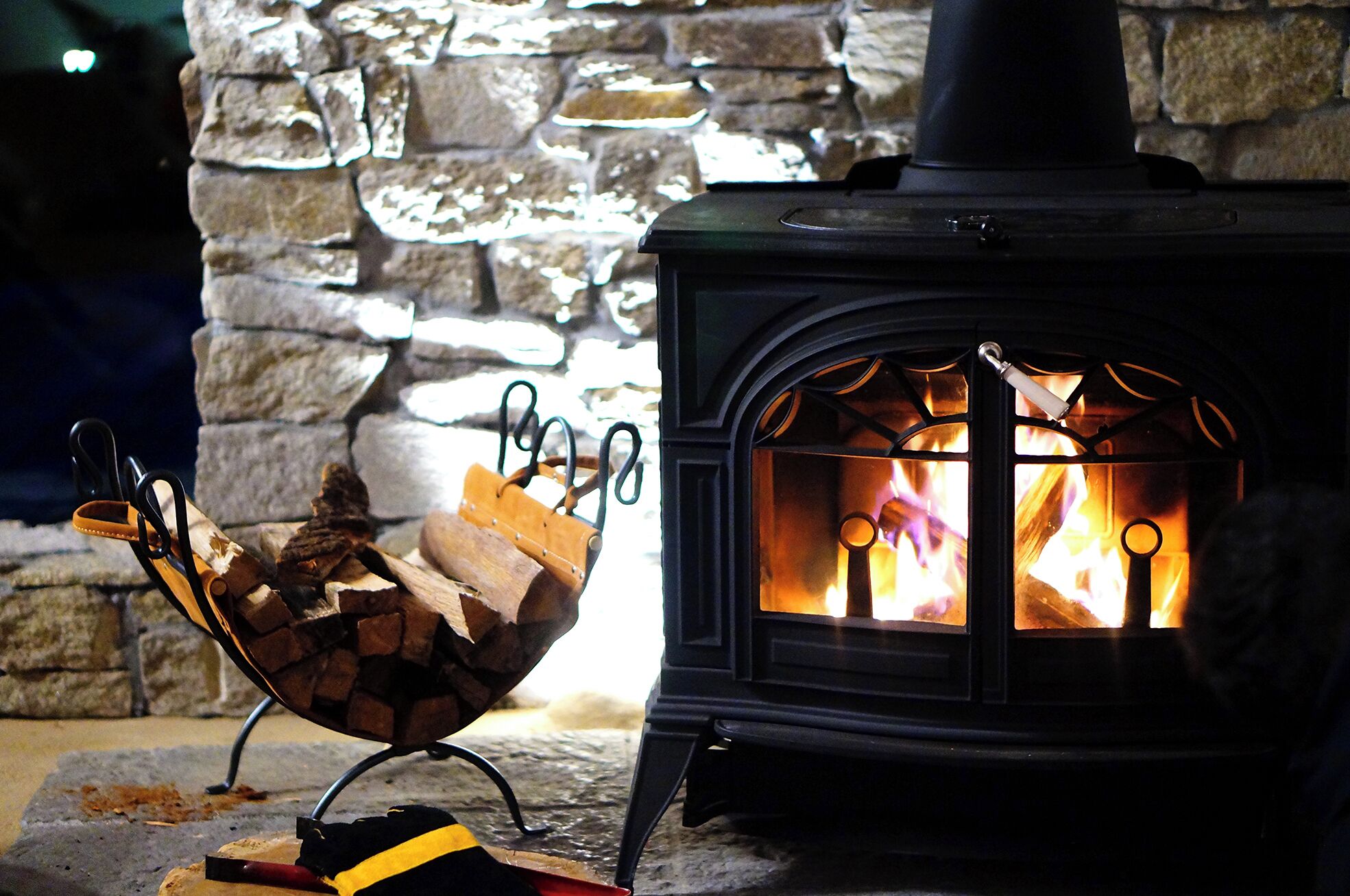 A wood-burning stove with a fire burning inside, set against a stone wall.