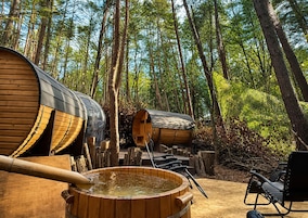 Un área boscosa con construcciones de madera, incluyendo una bañera grande de madera y un edificio cilíndrico, probablemente una sauna o jacuzzi.