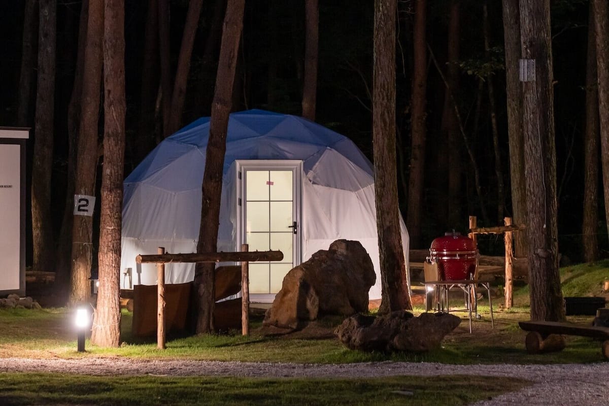 Une tente avec une porte, entourée d’arbres, et un barbecue situé à proximité.