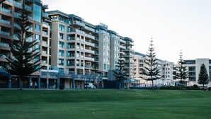 A modern multi-story residential building with balconies, a grassy courtyard, and a few pedestrians. - Sea the Views (Glenelg)