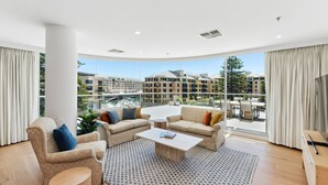 A spacious living room with a large window, a sofa set, a coffee table, and a view of a residential building. - Sea the Views (Glenelg)