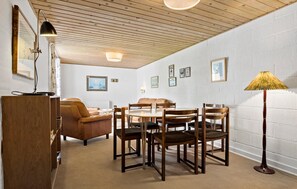 Ein gemütlicher Raum mit Holzbalkendecke, einem Esstisch mit Stühlen, einer Couch, einer Stehlampe und gerahmten Bildern an der Wand.
