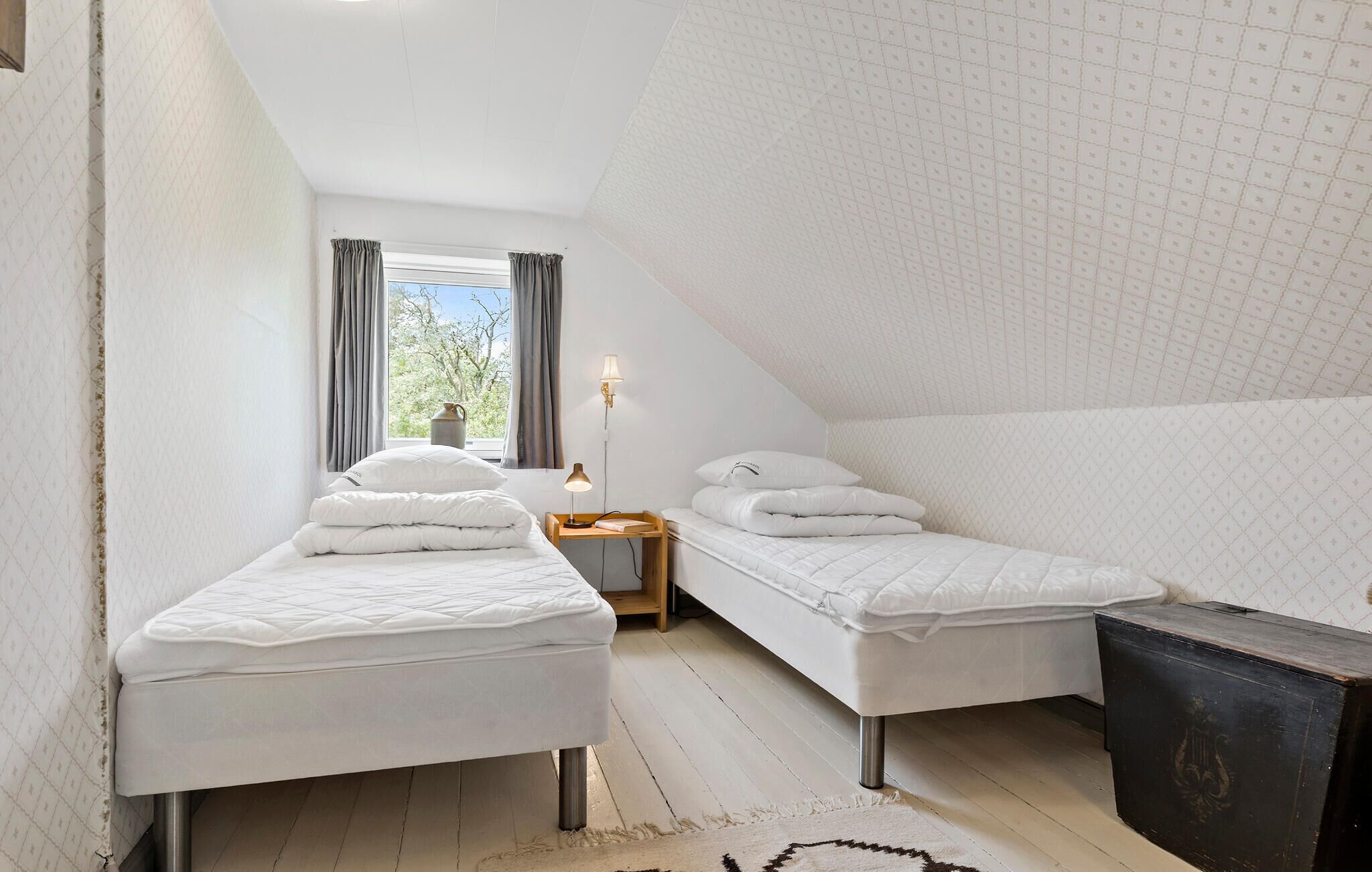 Een kleine, moderne slaapkamer met twee enkele bedden, een nachtkastje, een lamp en uitzicht op bomen door een raam.