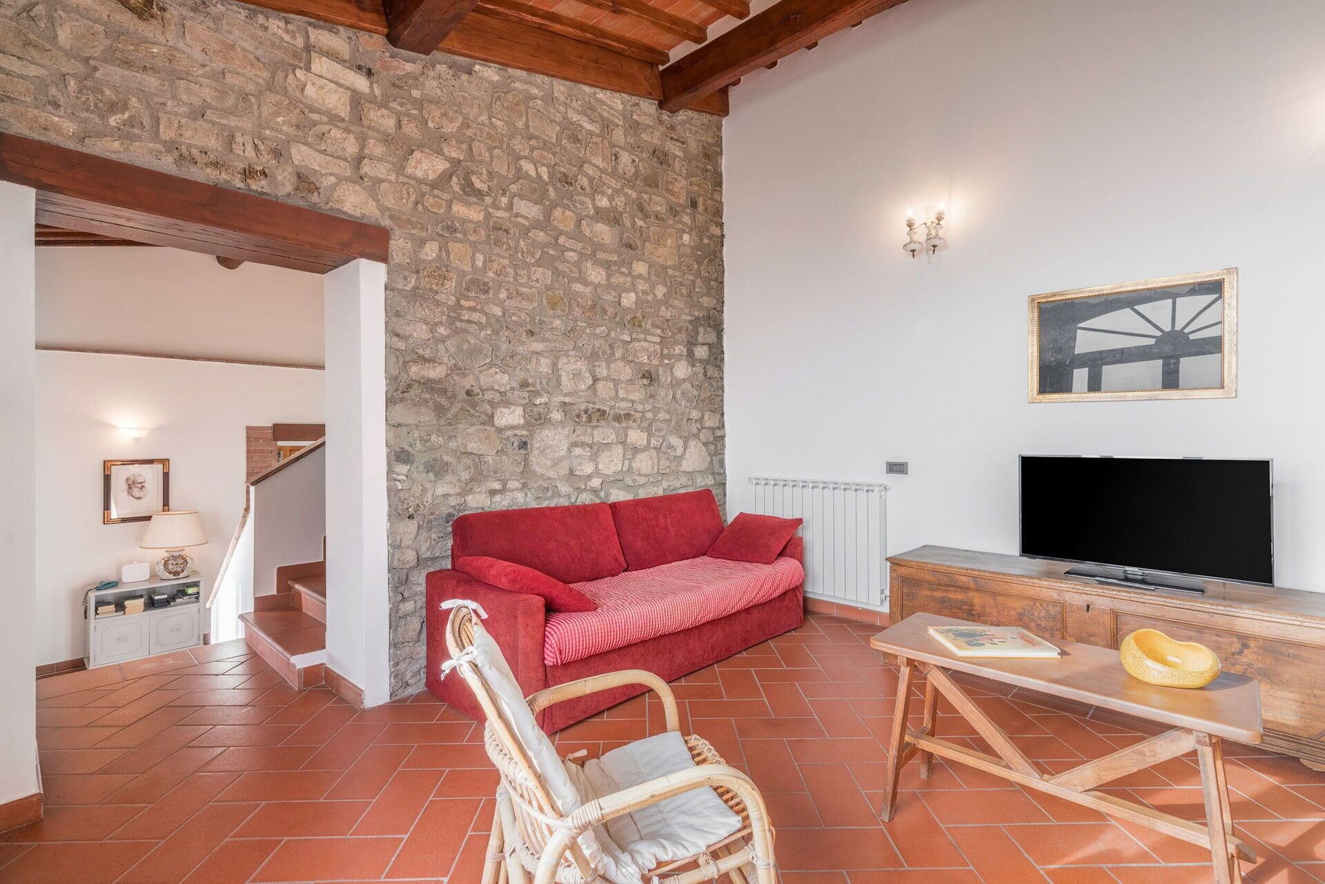 Un salon chaleureux avec un mur en pierre, un canapé rouge, un fauteuil en osier, une table en bois et un téléviseur.