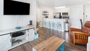 Un salon moderne avec un téléviseur à écran plat, une table basse en bois et un canapé en cuir. On aperçoit au fond une cuisine avec des meubles blancs et un coin bar.