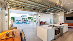 A modern kitchen with a large island, a dining area with a wooden table, and a view of an outdoor pool area. - Luxury 3bdr Villa with Private Pool, Oceanview and Hotel Amenities v4 (Providenciales)