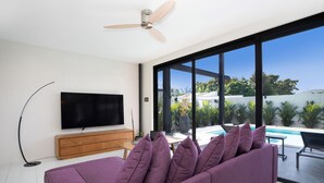 A modern living room with a large TV, purple sofa, wooden cabinet, and a view of a swimming pool and greenery. - Innovative 12m Eco Pool Villa Sunset 1 (Rawai)