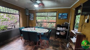 Een kamer met een ronde tafel, stoelen, een plafondventilator en een raam met uitzicht op groen.