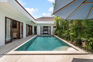 A rectangular swimming pool with a tiled deck, surrounded by a white building with glass doors and windows, and a covered area with a blue canopy. - Bamboo Damai Villa by Betterplace (Kerobokan)