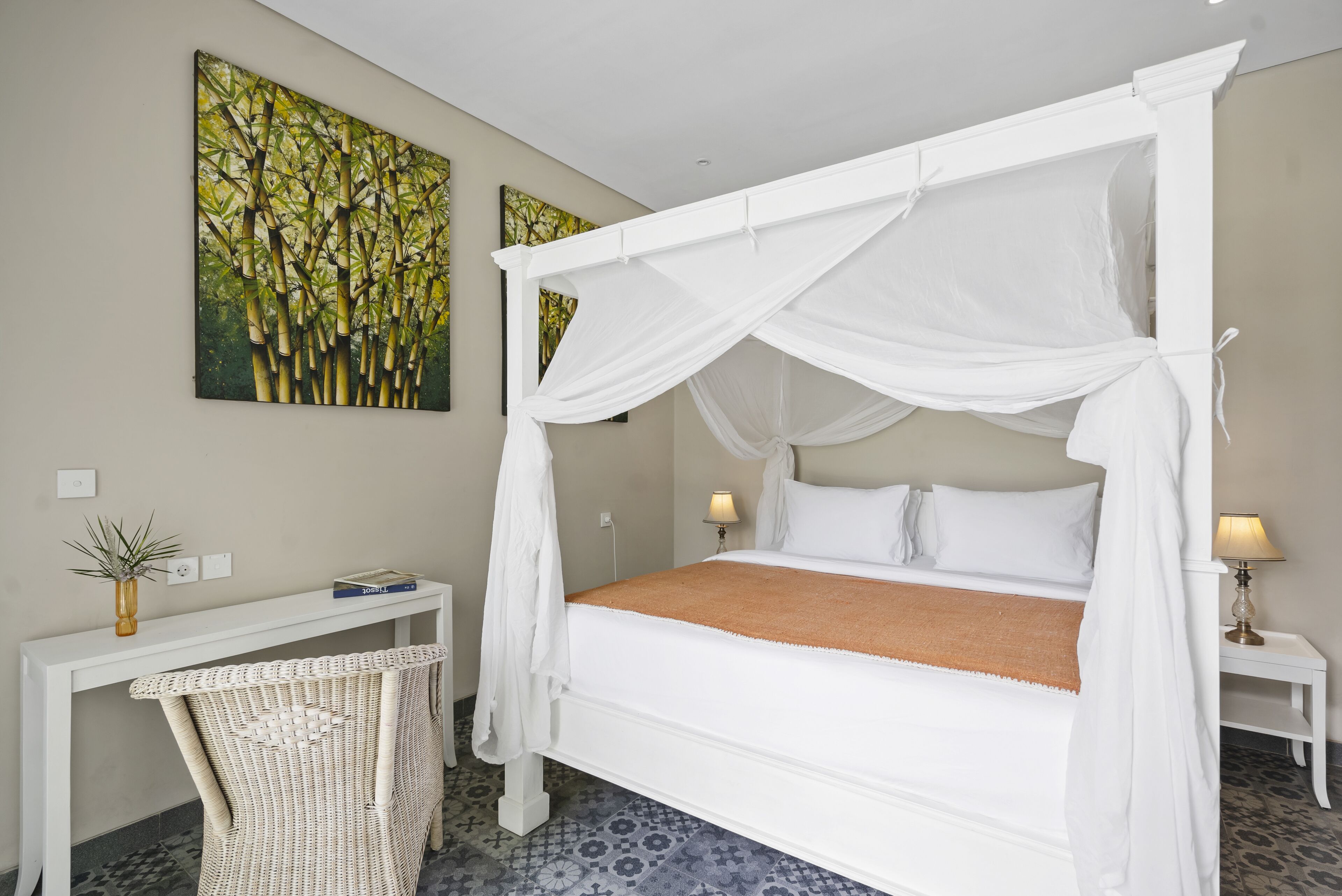 A four-poster bed with a white canopy, a bedside table with a lamp, a wicker chair, and a painting of bamboo on the wall.