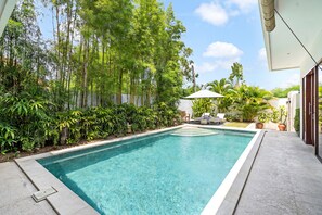 A rectangular swimming pool with clear blue water, surrounded by lush greenery and a white umbrella. - Bamboo Damai Villa by Betterplace (Kerobokan)