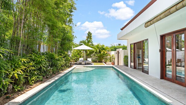 A rectangular swimming pool with clear blue water, surrounded by lush greenery and a modern house with large glass doors. - Bamboo Damai Villa by Betterplace (Kerobokan)