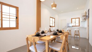 A dining area with a wooden table set for six, surrounded by chairs, with a kitchen in the background. - Japanese Style Villa Sunrise Garden B190 (Thep Krasattri)