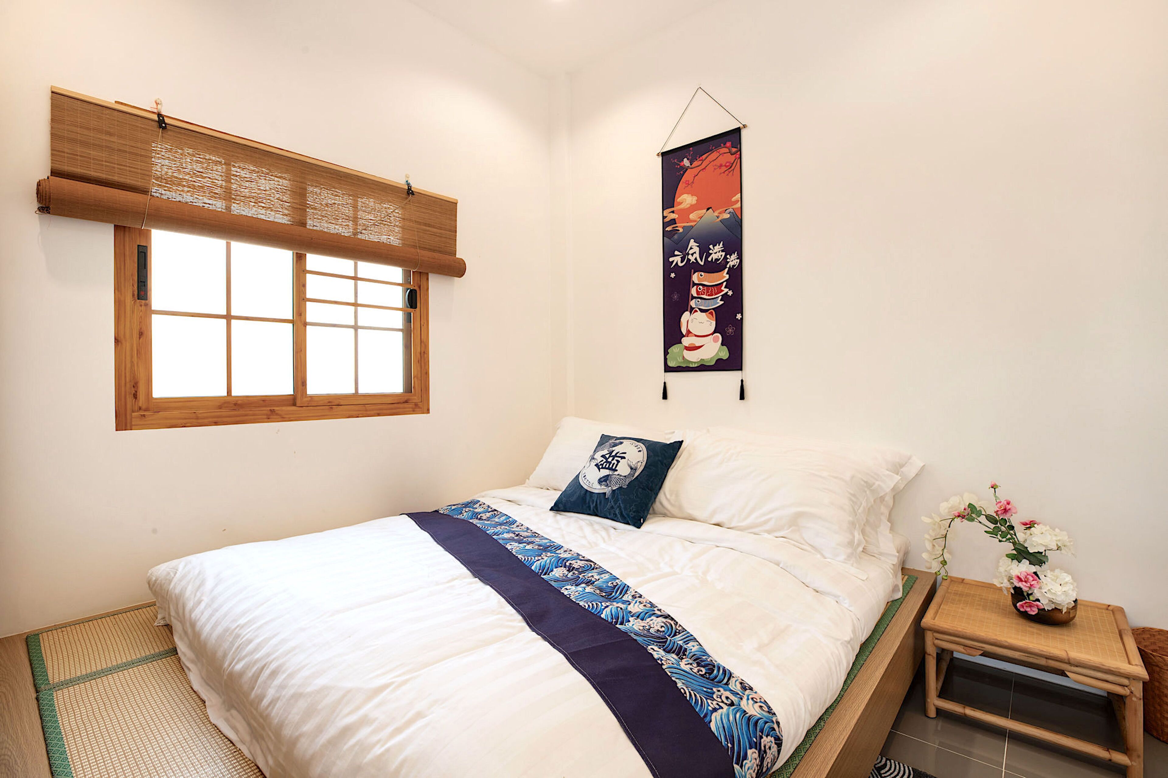 A neatly made bed with a blue and white bedspread, a wooden headboard, a window with a bamboo blind, and a wall hanging with Japanese characters.