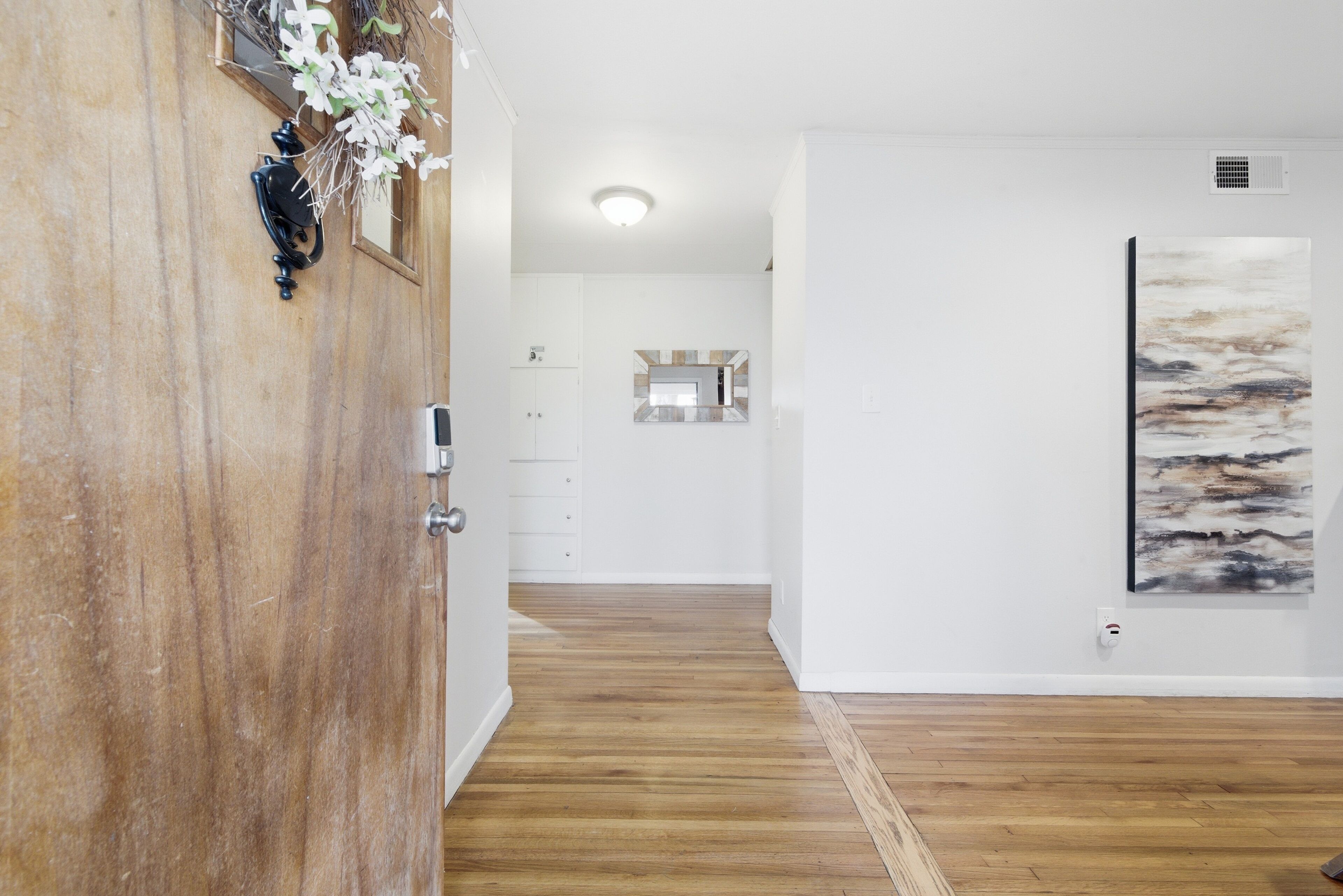 A hallway with wooden flooring, a white wall with a painting, and a kitchen area in the background.