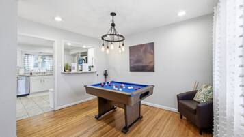 Une pièce avec une table de billard, un fauteuil sombre, un lustre et, au fond, une cuisine.