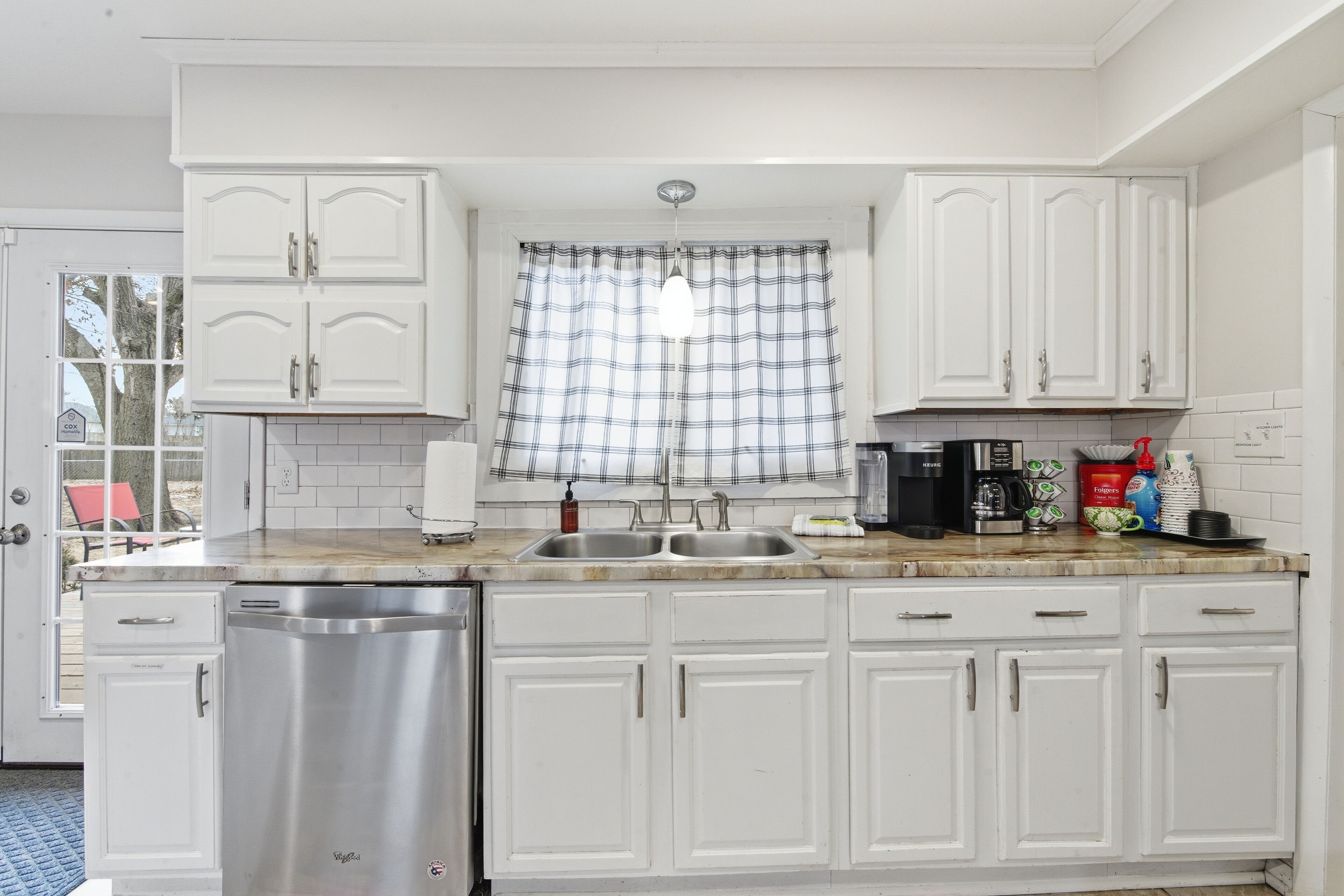 A kitchen with white cabinets, a stainless steel dishwasher, a double sink, and a window with plaid curtains.
