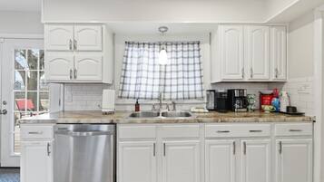 A kitchen with white cabinets, a stainless steel dishwasher, a double sink, and a window with plaid curtains.