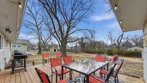 An outdoor patio with a glass table and red chairs, surrounded by a wooden deck and a large tree. - Near WSU:  Bumper Pool and Fire Pit! (Wichita)