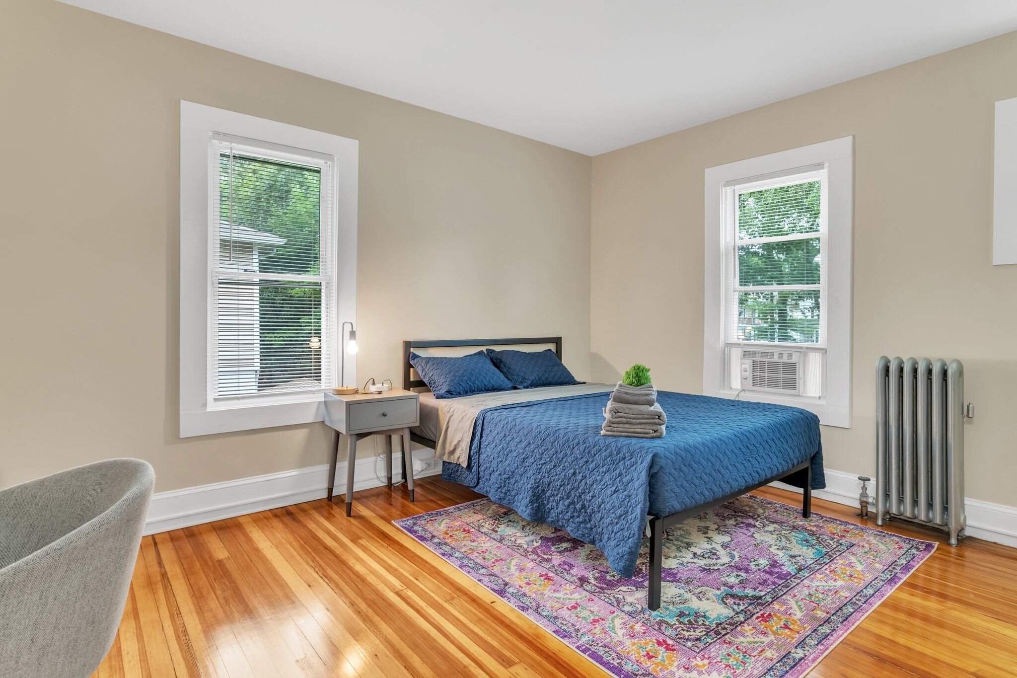 Quarto com cama, mesa de cabeceira, cadeira, radiador e duas janelas com persianas.