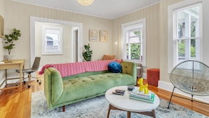 Un salon avec un canapé vert, une table basse ronde et un fauteuil. Il y a deux fenêtres équipées de stores, une plante et un luminaire suspendu.