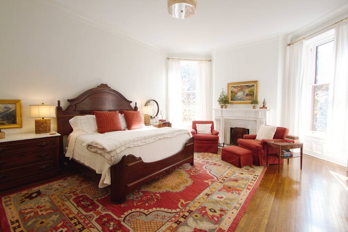 A spacious bedroom with a large bed, a red armchair, a small table, and a fireplace.