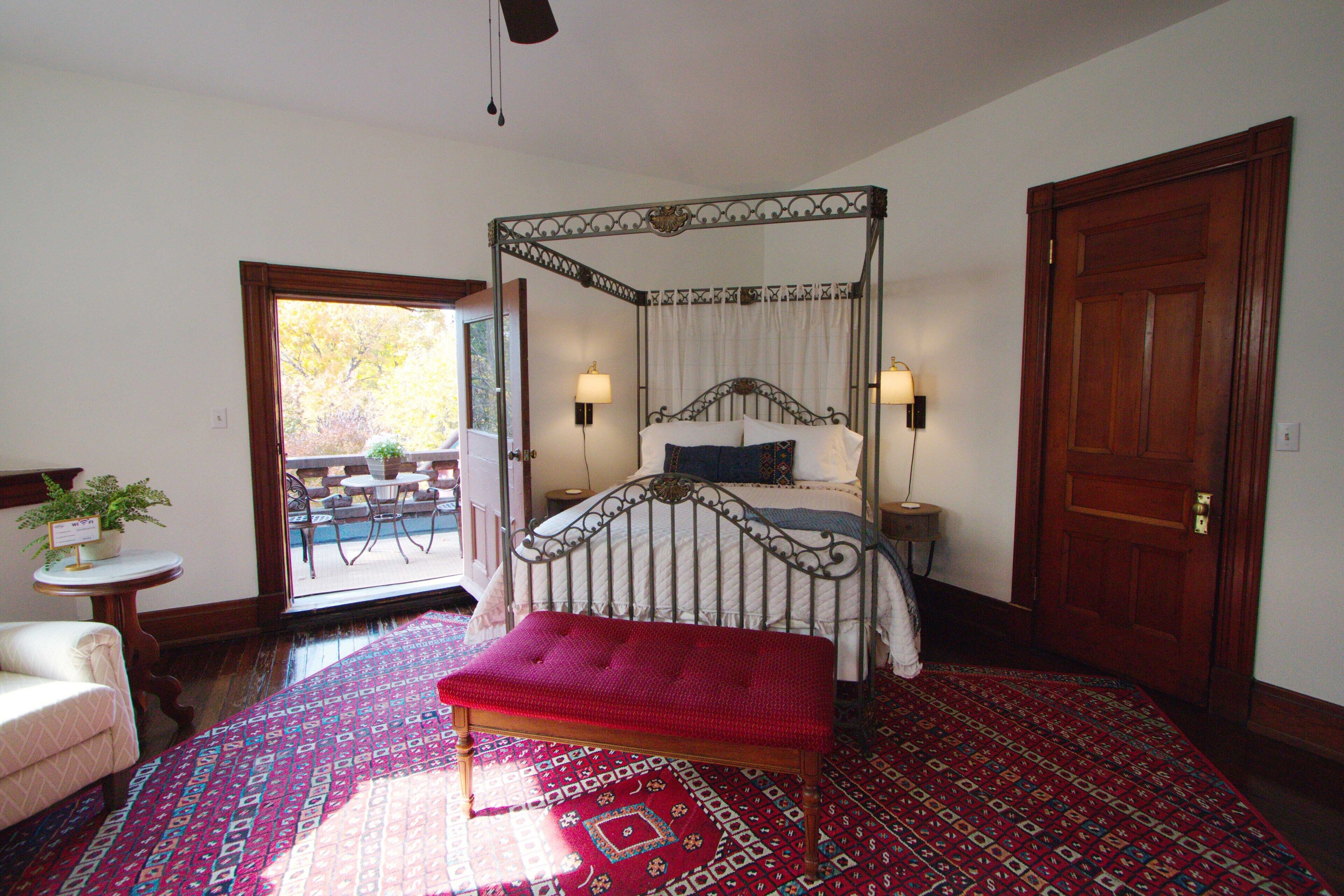 A four-poster bed with a red bench, a small table with a plant, and a view of the outdoors through a glass door.