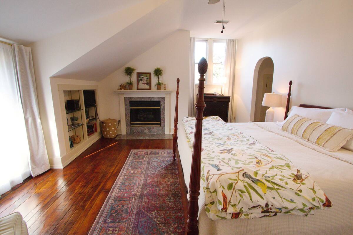 A bedroom with a four-poster bed, a fireplace, a large window, and a wooden floor.
