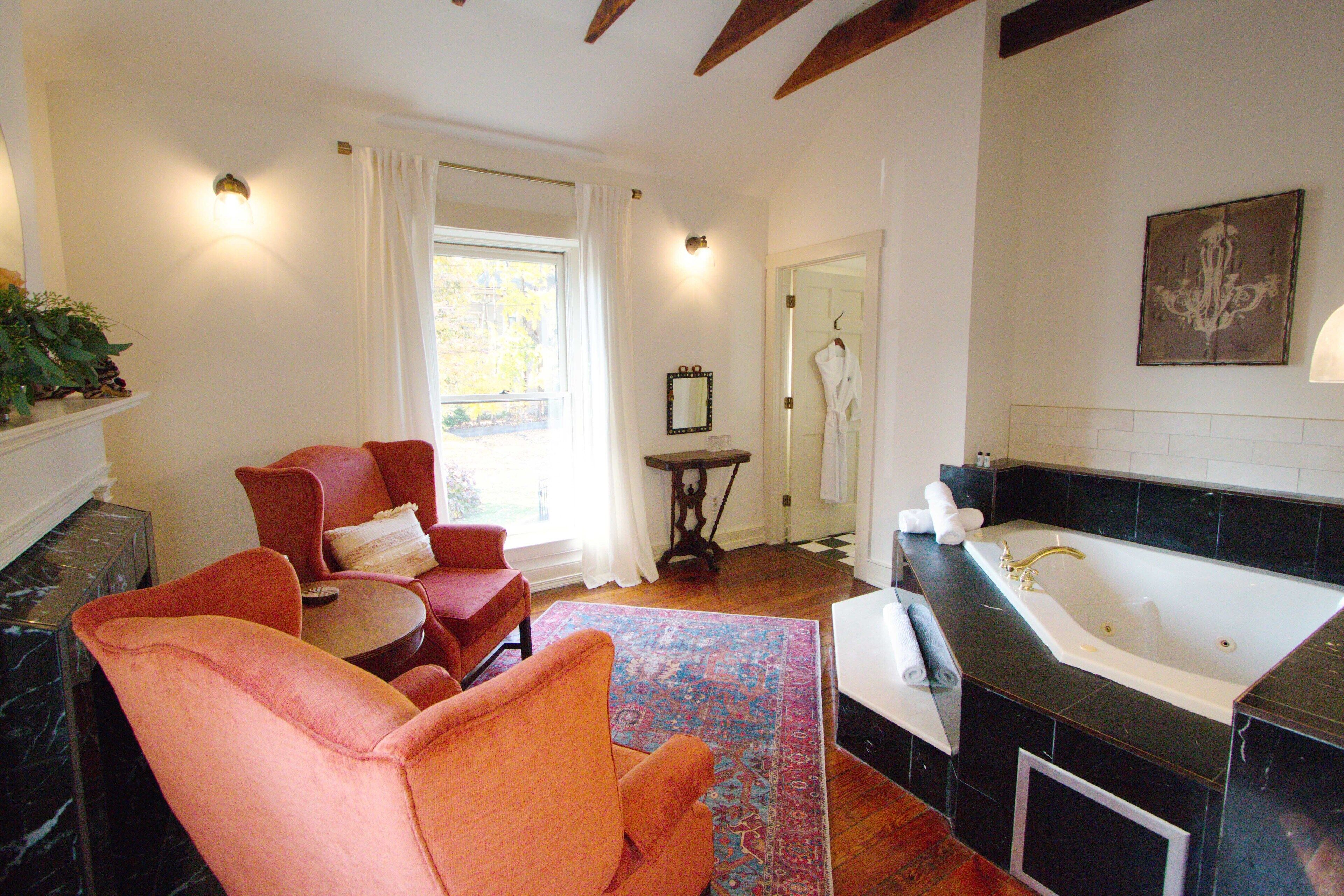 A room with a large window, a sitting area with chairs, a small table, and a bathroom with a bathtub.