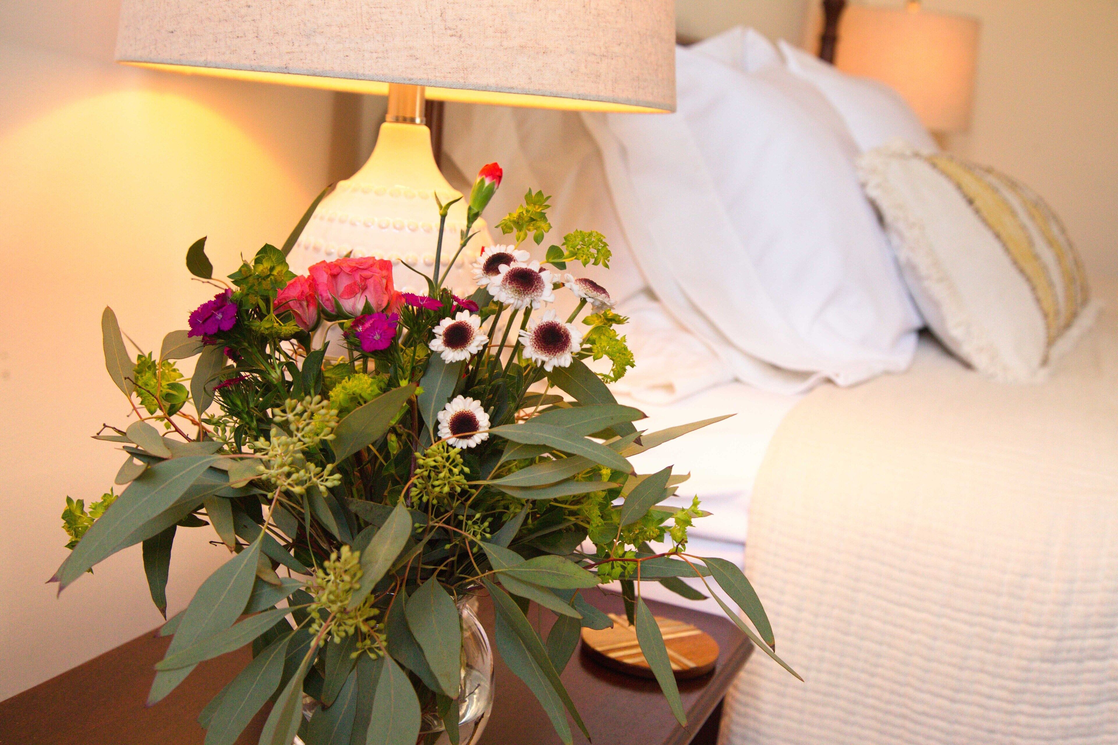 Une table de chevet avec un vase de fleurs, une lampe et un lit recouvert de linge de lit blanc.