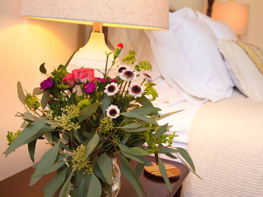 Une table de chevet avec un vase de fleurs, une lampe et un lit recouvert de linge de lit blanc.