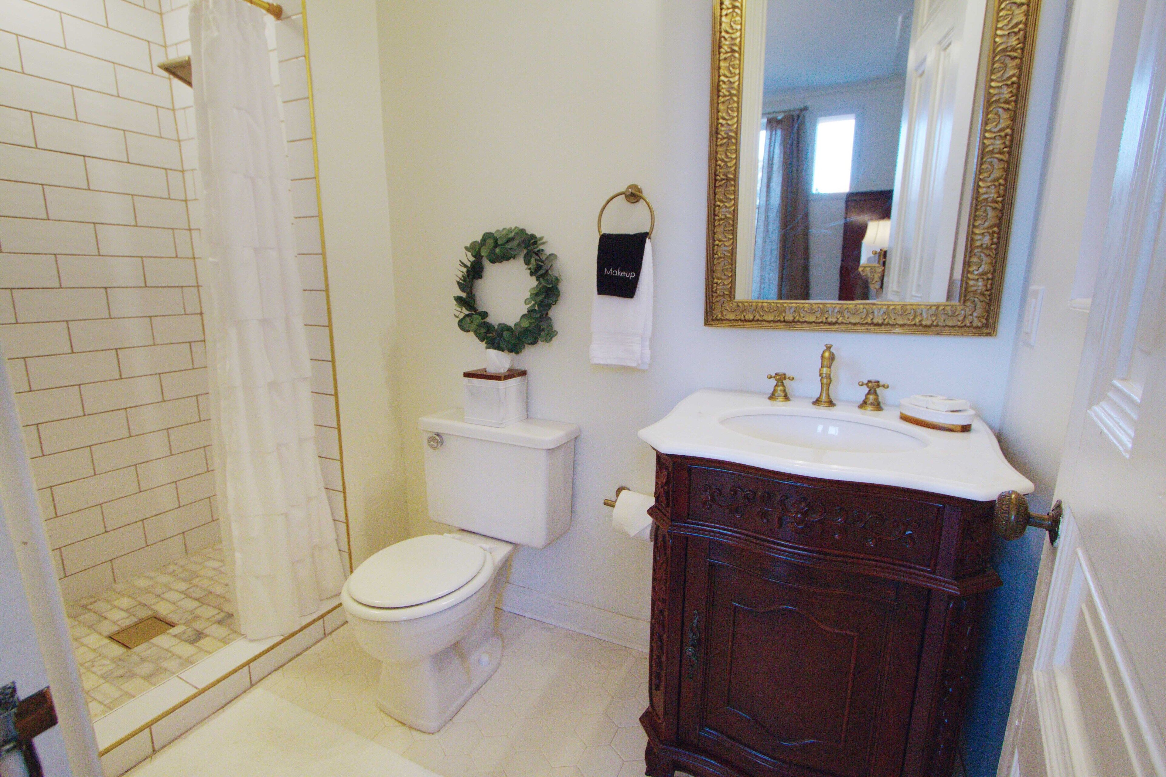 Une salle de bain avec des toilettes blanches, une douche avec un rideau blanc, un lavabo avec un robinet doré et un miroir avec un cadre doré.
