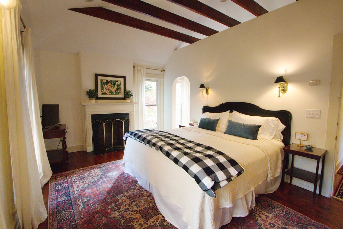 A bedroom with a large bed, a fireplace, a nightstand, and a window with curtains.