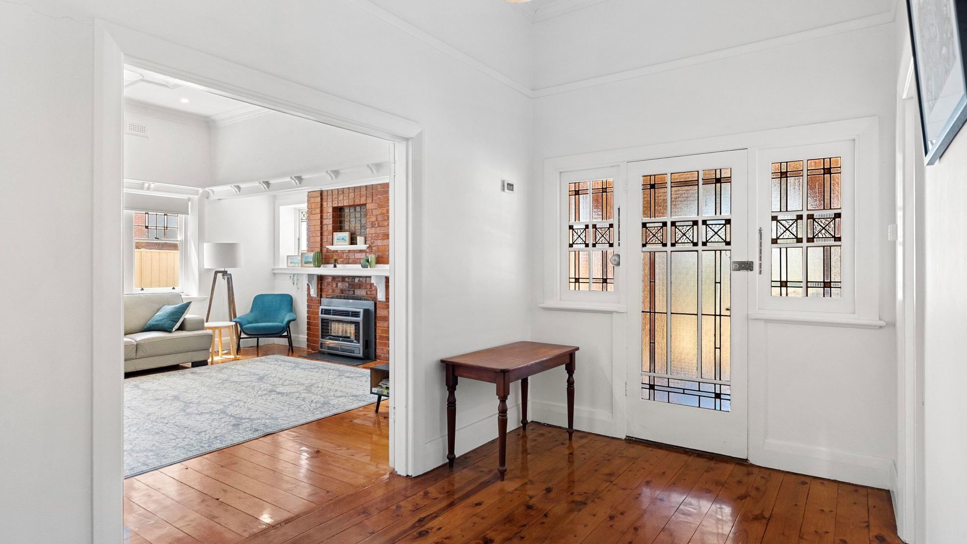 A spacious hallway with wooden flooring, a white door with stained glass panels, and a brick fireplace in the background.