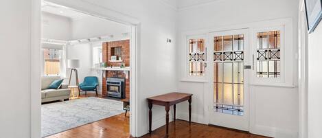 A spacious hallway with wooden flooring, a white door with stained glass panels, and a brick fireplace in the background.