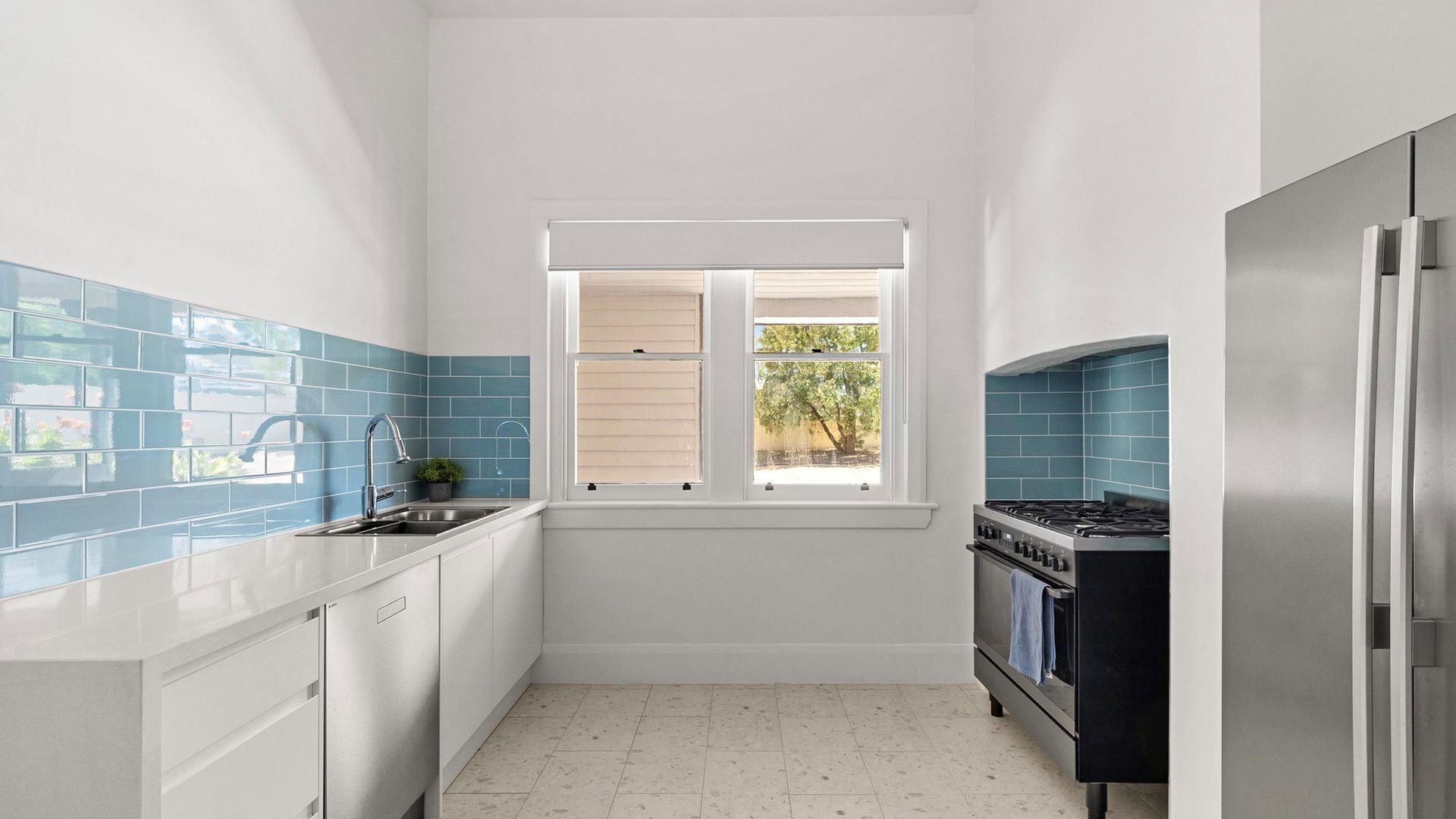 A compact kitchen with white cabinets, blue-tiled backsplash, a window with blinds, a stainless steel refrigerator, and a black stove.